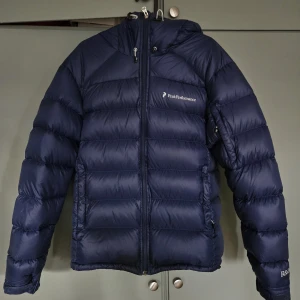 Mörkblå dunjacka Peak Performance S - Säljer en mörkblå dunjacka från Peak Performance i storlek S. Jackan har huva, dragkedja framtill och är tillverkad i lätt och slitstark Pertex Microlight. Perfekt för kalla dagar och har en clean, sportig look.