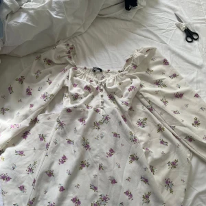 Blommig vit klänning Gina Tricot XS - Supersöt vit klänning från Gina Tricot i storlek XS med lila och gröna blommor. Klänningen har långärmade puffärmar, smock vid halsen och knappar framtill. Säljer pga den inte kommer till användning❤️