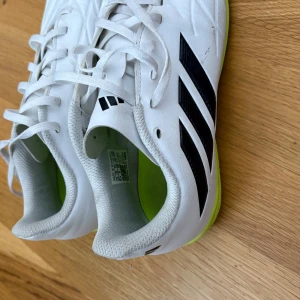Vita adidas fotbollsskor strl 38 - Snygga vita adidas fotbollsskor med svarta ränder och neongul sula. Skorna har snörning och är tillverkade i syntetmaterial. Perfekta för inomhusspel med sin platta, greppvänliga sula och sportig look.