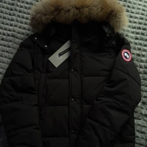 Svart dunjacka från Canada Goose - Säljer en svart dunjacka från Canada Goose med stor fluffig päls på huvan och klassisk logga på ärmen. Jackan är quiltad och har dragkedja samt knappar framtill. Perfekt för kalla vinterdagar och riktigt snygg streetstil.