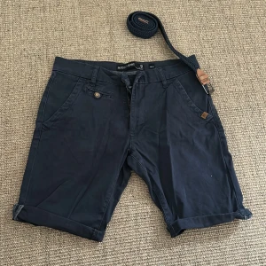 Mörkblå shorts från INDICODE JEANS - Snygga mörkblå shorts från INDICODE JEANS med klassisk design, uppvikta benslut och medföljande flätat bälte. Shortsen har flera fickor, knapp- och dragkedjestängning samt detaljer i brunt läder. Perfekta för sommardagar och chill häng.