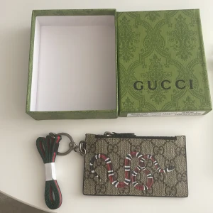Gucci plånbok med orm och GG-mönster - Gucci plånbok i beige och svart med klassiskt GG-mönster och en cool röd, vit och svart orm på framsidan. Baksidan har svarta kortfack i skinn och dragkedja upptill. Nyckelring och grön-röd rem medföljer. Kommer med original ask och dustbag.
