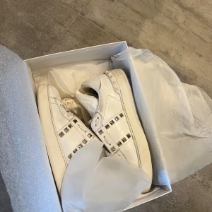 Valentino Rockstud vita sneakers - Snygga vita Valentino Rockstud sneakers i skinn med ikoniska silverfärgade nitar längs sidorna. Klassisk rund tå, platt sula och vita snören. Perfekta för dig som vill ha en lyxig och edgy touch till din outfit.   Ta kontakt for frakt