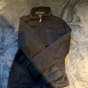 Svart vindjacka från Peak Performance - Svart vindjacka från Peak Performance med klassisk krage och dragkedja framtill. Jackan har ett rutigt innerfoder i vitt och blått, samt diskret logga på bröstet. Perfekt för dig som vill ha en stilren och funktionell jacka till vardags.