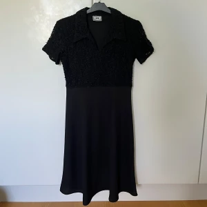 Svart klänning med spets från Joy - Svart klänning från Joy med krage och korta ärmar. Överdelen har ett mönster i spets och nederdelen är slät.