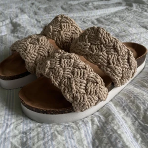 Beige sandaler med flätade band - Snygga sandaler från Exani med två breda, flätade band i beige. Sulan är vit och har en vågig struktur undertill för extra grepp. Perfekta för varma dagar och ger en chill boho-vibe till din outfit.