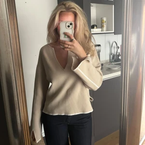 Beige stickad tröja med v-ringning - Säljer en beige stickad tröja med v-ringning och långa ärmar. Tröjan har en loose passform, vita detaljer längs ärmar och nederkant samt slits vid ärmsluten. Perfekt för dig som gillar en clean och avslappnad stil.