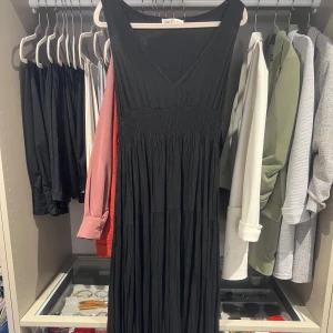 Svart långklänning med v-ringning - En svart långklänning med v-ringning och smockad midja. Klänningen är ärmlös och har ett luftigt, fallande tyg. Riktigt fin! 