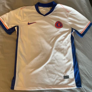 Chelsea FC vit Nike fotbollströja S - Chelsea FC fotbollströja från Nike i ljusrosa med blå detaljer och röda inslag. Kortärmad med blå V-ringad krage och blå/röda ränder på ärmsluten. Tillverkad i lätt Dri-FIT material som andas, perfekt för träning eller match. Chelsea-logga och Nike Swoosh på bröstet. Stor för att vara S är som M