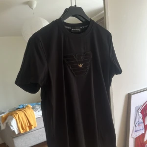 Svart t-shirt Emporio Armani M - Svart t-shirt från Emporio Armani med ikoniskt broderat örnlogo i svart och guld på bröstet samt vit logga på ärmen. Klassisk rund hals och korta ärmar. Mjuk och skön bomullskvalitet, perfekt för dig som gillar stilrena designer med lyxig känsla.