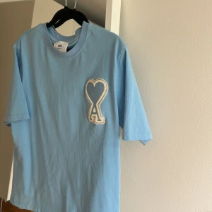 Blå Ami t-shirt med hjärtdetalj - Snygg ljusblå t-shirt från Ami med ett stort broderat hjärta och bokstaven A på bröstet. Klassisk rund hals och korta ärmar. Tillverkad i mjuk bomull som känns skön mot huden. Perfekt för dig som gillar stilrena plagg med en twist.