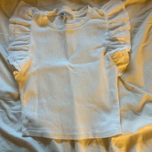 Vit volangtopp från Zara - Supersöt vit topp från Zara med ribbad struktur och volangdetaljer längs axlarna. Modellen är ärmlös och har en rund halsringning. 