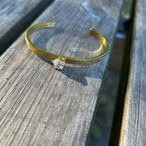 Guldigt armband med kristall - Snyggt och stilrent armband i guldton med en glittrande kristall som detalj framtill. Öppen design som gör det enkelt att ta på och av. Perfekt för dig som gillar minimalistiska smycken med en touch av bling.