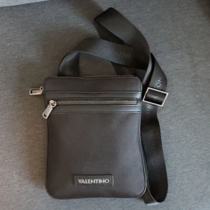 Svart axelväska från Valentino - Snygg svart axelväska från Valentino med justerbart band där loggan är tryckt. Väskan har två dragkedjefack, en framtill och en baktill, och är tillverkad i slitstark syntet med detaljer i skinnimitation. Perfekt storlek för mobil, plånbok och nycklar.