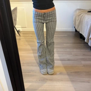 Randiga lågmidjade bootcut byxor - Säljer ett par grå och vita randiga byxor med låg midja och bootcut-snitt. Byxorna har en klassisk dragkedja och knapp framtill. Snygg passform som sitter tajt upptill och släpper ut vid benen. Perfekta för dig som gillar retrovibbar och vill sticka ut.