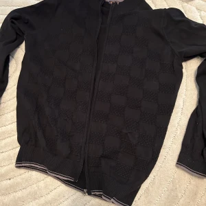 Svart kofta med rutigt mönster - Svart kofta med dragkedja och diskret rutigt mönster framtill. Tröjan har ribbade muddar och krage med tunna kontrastlinjer i vitt och grått. Perfekt för dig som gillar stilrena och klassiska plagg med en twist.