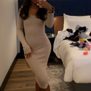 Beige ribbad långklänning - Snygg beige långklänning med ribbad struktur och v-ringad hals. Klänningen har långärmade ärmar med slits vid ärmslut och sitter tight mot kroppen för en smickrande siluett. Perfekt för dig som gillar stilrena och enkla plagg med en modern touch.