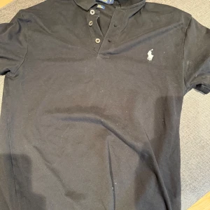 Svart Polo Ralph Lauren Pike - Svart pike från Polo Ralph Lauren i slim fit med stretch mesh-material. Klassisk pikékrage och tre knappar framtill, samt den ikoniska broderade loggan på bröstet. Perfekt för dig som gillar stilren och enkel design.