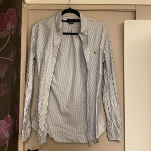 Randig skjorta från Polo Ralph Lauren - Snygg slim fit skjorta från Polo Ralph Lauren i vit och ljusblå randigt mönster. Klassisk krage, långa ärmar och knappar framtill. Broderad logga på bröstet. Perfekt för dig som gillar stilrena och tidlösa plagg.
