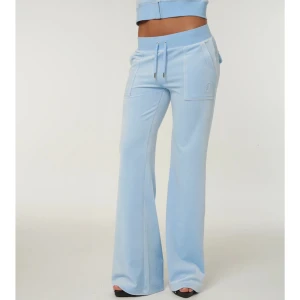 Ljusblå velourbyxor Juicy Couture - Supermjuka ljusblå velourbyxor från Juicy Couture med bootcut passform. Byxorna har bred resår i midjan med snörning, stora fickor fram och bak med lock och metallknappar. Perfekta för en chill och trendig look.