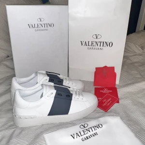 Valentino Garavani Open sneakers vit/blå - Snygga Valentino Garavani Open sneakers i vitt skinn med bred mörkblå rem över vristen. Klassisk låg modell med rund tå, vita snören och ikoniska nitar på hälen. Perfekta för dig som vill ha en clean och lyxig look. 0bs 1nte 0rg1nal!