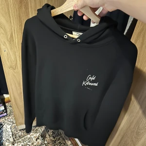 Maison kitsune hoodie - Snygg svart hoodie från Café Kitsuné (maison kitsune)  med vit logga på bröstet och ryggen. Perfekt för en stilren och avslappnad look. Den har en klassisk huva med dragsko och långa ärmar. 