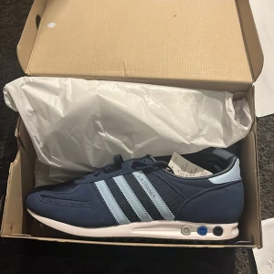 Adidas LA Trainer marinblå sneakers - Säljer ett par Adidas LA Trainer sneakers st 44 i marinblått med ljusblå ränder och detaljer. Skorna har klassisk snörning, vit sula med de ikoniska pluggarna i blått och grått samt ovandel i mocka och mesh. Perfekta för dig som gillar retrostil och sportig vibe. Helt nya i kartong säljer endast då de är för stora och inte går att skicka tillbaka.