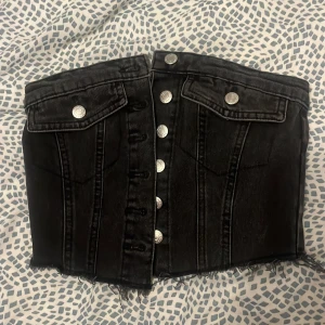 Svart jeanskorsett med knappar -  svart korsettopp i jeansmaterial med silvriga knappar framtill och två fickor med lock. Toppen har råa, fransiga kanter nertill och smockad rygg för extra stretch