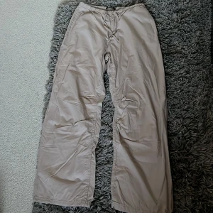 Beiga parachute byxor från Gina Tricot - Säljer ett par beiga wide byxor från Gina Tricot i storlek M. Byxorna har resår i midjan, snörning vid benslut och en bakficka med lock. Perfekta för en chill och avslappnad stil. Materialet känns lätt och luftigt.