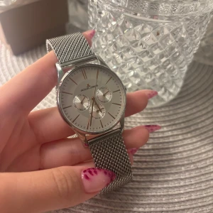 Jacques Lemans silverfärgad klocka - Stilren klocka från Jacques Lemans i silver med mesh-armband i rostfritt stål. Urtavlan är rund och har två mindre subdials för dag och datum. Klassisk design med minimalistiska index och diskret logga på urtavlan. Perfekt accessoar för en clean look. Går ej att köpa längre, nypris ca 1900kr 