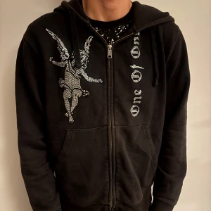 One of one tröja  - Svart hoodie med dragkedja och huva. På framsidan finns en stor ängel i silvriga stenar och texten 'one of one' i liknande stil. Tröjan har känguruficka och långa ärmar. Perfekt för dig som gillar streetwear med unika detaljer.