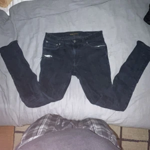 Nudie jeans - Säljer ett par mörkblå skinny jeans från Nudie med klassiska bakfickor och broderad våg. Jeansen har en snygg sliten detalj på ena låret och är tillverkade i mjuk bomullsdenim. Perfekta för dig som gillar en tajt passform och stilren look.