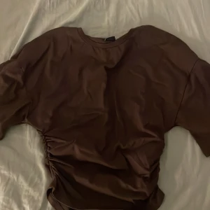 Brun t-shirt med scrunch i sidan från Gina - Snygg brun t-shirt med korta ärmar och cool snörningsdetalj på ena sidan som ger en unik look. Rund halsringning och loose passform, perfekt för dig som gillar stilrena plagg med en twist. Materialet känns mjukt och skönt mot huden. Storlek S, aldrig använd.