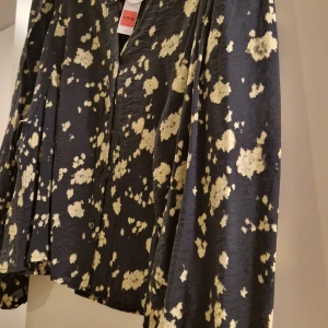 Svart blommig blus från Lindex - Supersnygg svart blus från Lindex med ljusgult blommönster och ballongärmar. Blusen har knappar framtill och en lätt glansig yta som ger en cool vibe. Perfekt för dig som vill sticka ut lite extra med ett unikt print och voluminösa ärmar. Obs! Storleken är ganska liten,L passar som M om man har stora bröst. Helt slut på online. 