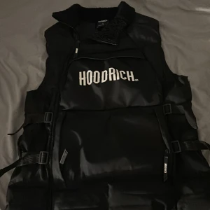 Svart dunväst från Hoodrich - Snygg svart dunväst från Hoodrich med stor vit logga framtill. Västen har hög krage med mjukt foder, justerbara spännen i sidorna och dragkedja. Perfekt för lager-på-lager 