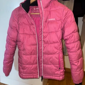 Rosa pufferjacka från Everest 146 - Snygg rosa pufferjacka från Everest med huva och dragkedja. Jackan är quiltad och har två fickor framtill. Perfekt för kalla dagar och riktigt skön att ha på sig. Materialet är 100% polyester och jackan har en sportig vibe.