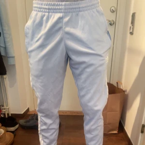 Ljusblå Adidas mjukisbyxor - Säljer ett par ljusblå mjukisbyxor från Adidas med klassiska vita ränder längs sidorna och logga på vänster ben. Byxorna har elastisk midja och smal passform som sitter snyggt. Perfekta för chill eller träning, riktigt sköna och stilrena.
