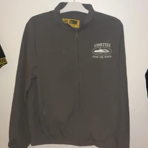 Corteiz Windbreaker - Grå vindjacka från Corteiz Litework med broderad logga och text på bröstet samt ärmen. Jackan har dragkedja framtill, ståkrage och praktiska fickor. Perfekt för dig som gillar streetwear och vill ha en clean look.