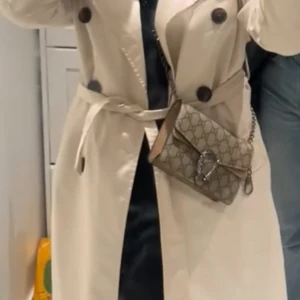 Beige trenchcoat från Pull&Bear XS -  beige trenchcoat från Pull&Bear med  krage, stora svarta knappar och bälte i midjan.  Lätt material som passar bra till lager-på-lager. 