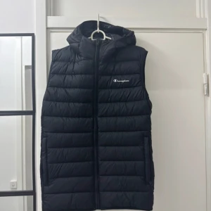 Svart dunväst från Champion - Svart dunväst från Champion med huva och dragkedja framtill. Västen har quiltad design, logga på bröstet och längs sidorna. Perfekt för lager-på-lager och streetwear-vibe. Snygg och enkel att matcha med hoodies eller sweatshirts.