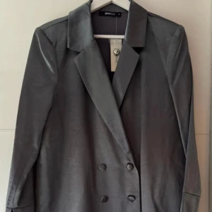 Ny. Blazer/klänning från Ginatricot  - Kan användas både som blazer eller klänning. Storlek 40, bröst 104cm, längd 83cm, viskos 58%, färg silver med lyster. Nypris 699kr