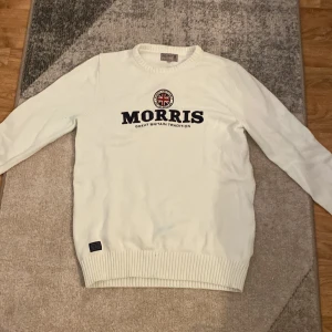 Vit stickad tröja från Morris - Snygg vit stickad tröja från Morris med broderad logga och brittisk flagga på bröstet. Rund hals och ribbade muddar vid ärmslut och nederkant. Perfekt för dig som gillar klassisk stil med en modern twist.