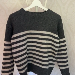 Randig grå stickad tröja - Mysig stickad tröja i mörkgrått med breda vita ränder över bröstet och ärmarna. Tröjan har en klassisk rund halsringning och långärmad passform, perfekt för kyliga dagar. Enkel och stilren design som funkar till det mesta.