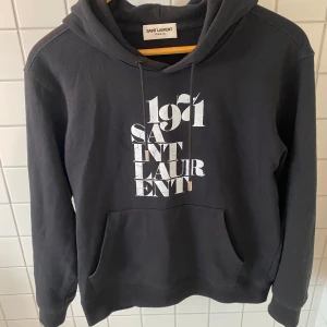 Svart hoodie från Saint Laurent - Svart hoodie från Saint Laurent med vit tryckt text på bröstet. Klassisk huva med snörning och stor magficka framtill. Mjuk bomullskvalitet och långärmad passform, perfekt för vintern! OBS köpt på plick så är inte hundra på om den är äkta.