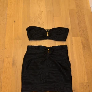 Svart bikini med gulddetalj - Snygg svart bikini med bandeau-topp och matchande underdel. Båda delarna har en cool guldfärgad ring i mitten och vågiga strukturer i tyget som ger en unik look. Perfekt för stranden eller poolen, riktigt trendig stil.