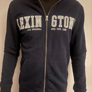 Mörkblå hoodie från Lexington - Snygg mörkblå hoodie från Lexington med dragkedja och stora vita bokstäver framtill. Tröjan har känguruficka och ribbade muddar. Perfekt för en chill och sportig look. Tillverkad i mjuk bomull för extra komfort.