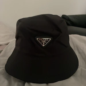 Prada hat bucket - Säljer en svart bucket hat från Prada med ikonisk metalltriangel framtill. Hatten har en stilren och modern look, tillverkad i slitstark polyester med snyggt foder. Perfekt för dig som vill ha en lyxig accessoar med street vibe och designerstil.