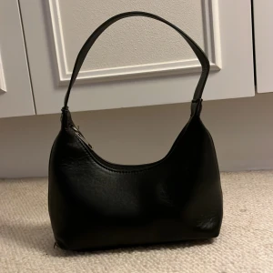 Svart axelväska i faux skinn - Snygg svart axelväska i faux skinn med minimalistisk design och mjuka former. Väskan har ett smidigt handtag och stängs med dragkedja upptill. Perfekt storlek för att bära med sig det viktigaste och passar till många olika outfits.