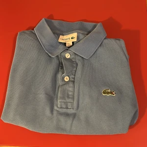 Ljusblå polo  från Lacoste - Säljer en ljusblå polo från Lacoste i slim fit-modell. Klassisk design med krage, knappar och det ikoniska krokodilmärket broderat på bröstet. Tröjan är i bomull och har korta ärmar, perfekt för en fräsch och stilren look.