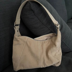 Beige axelväska i mocka - Snygg beige axelväska i mjuk mocka med bred axelrem i matchande färg. Väskan har silverfärgad dragkedja upptill och dekorativa sömmar framtill. Perfekt storlek för det viktigaste och enkel att slänga över axeln.